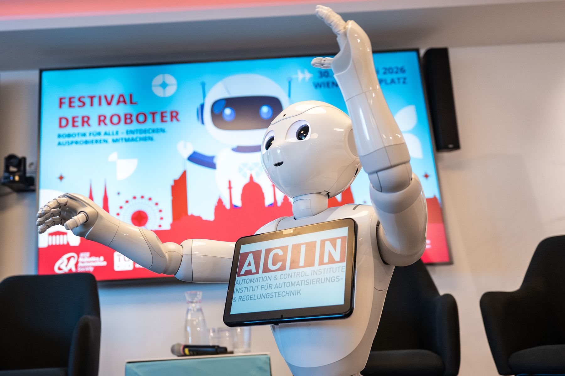 Kopf, Arme und Oberkörper eines kleinen weißen Roboters mit Tablet auf der Brust vor einem Bildschirm