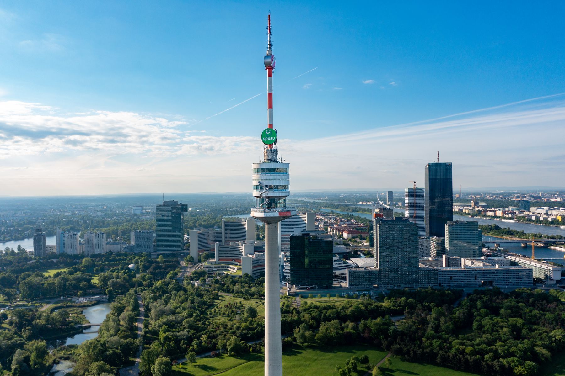 Blick aus der Luft auf den Donauturm in Wien mit rot-weißer Antennenspitze, umgeben von grünen Parkanlagen, moderner Hochhausarchitektur und der Donau unter blauem Himmel.