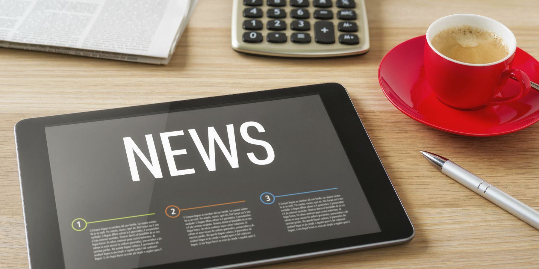 Auf einem Holztisch liegen ein Tablet mit schwarzen Hintergrund mit der Anzeige ‚News‘, ein Taschenrechner, Zeitungen und eine rote Kaffeetasse mit Kaffee.
