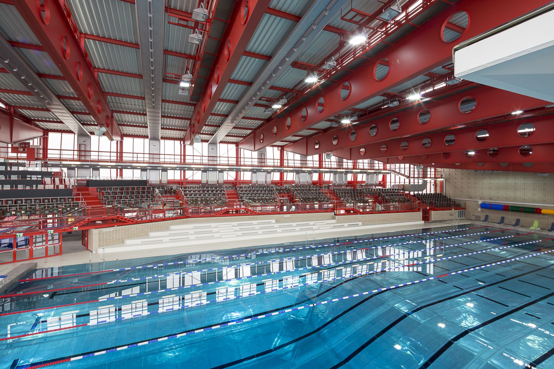 Modernes Hallenbad mit mehreren Schwimmbahnen und klarem Wasser. Im Hintergrund eine lange Tribüne mit grauen Sitzreihen. Rote Stahlträger und große Glasfronten prägen die helle, lichtdurchflutete Architektur.