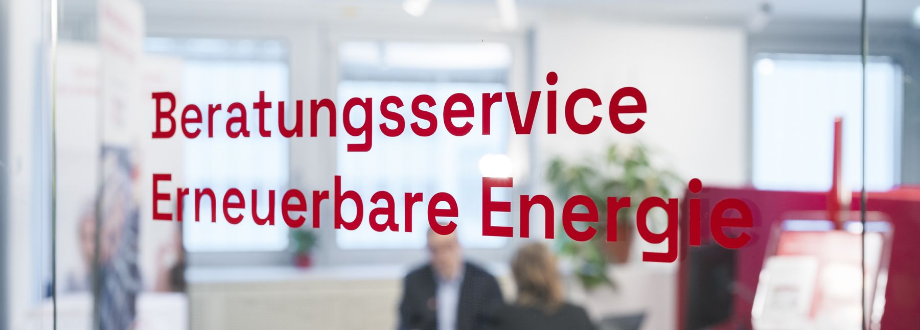 Durch eine Glasscheibe mit der roten Aufschrift „Beratungsservice Erneuerbare Energie“ sind zwei Personen unscharf bei einem Gespräch an einem Schreibtisch zu sehen.