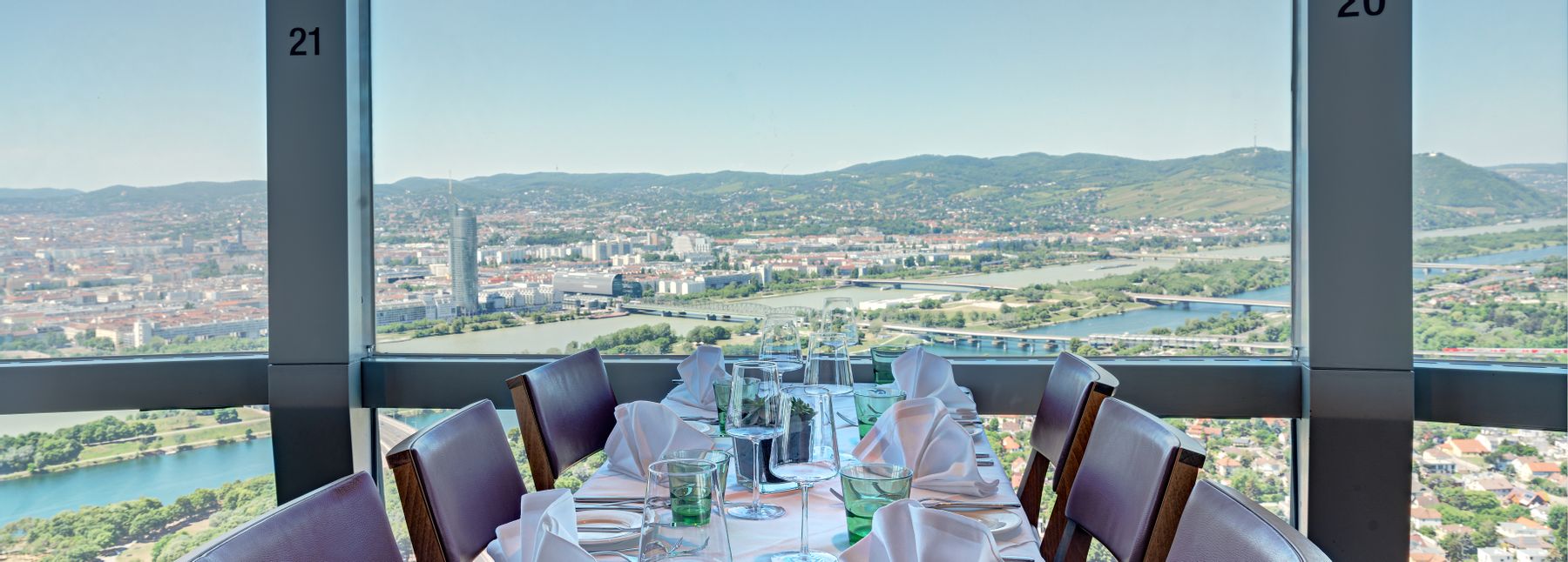 Gedeckter Tisch im Turmrestaurant des Donauturms in Wien mit weißer Tischwäsche, Gläsern und Servietten sowie Panoramablick über die Donau und die Wiener Stadtlandschaft.