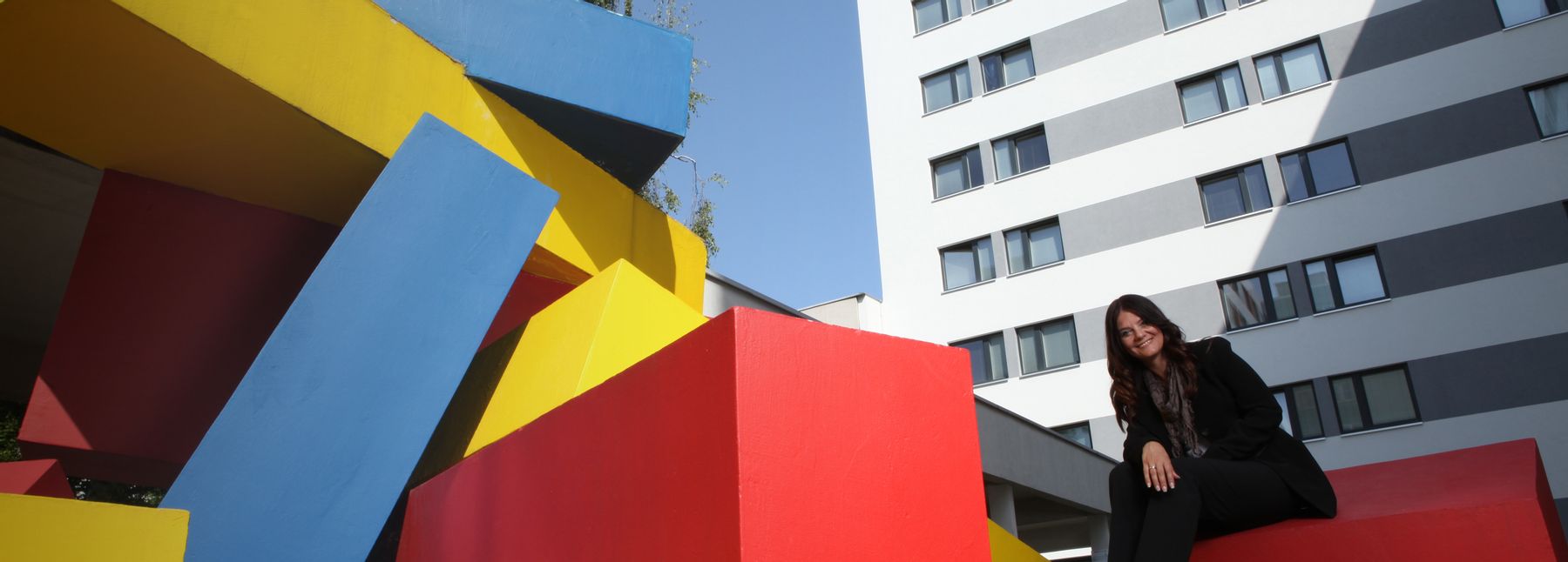 Außenansicht der Viennabase19, ein kastenförmiges Gebäude; im Vordergrund steht eine Kunstinstallation aus gelben, roten und blauen Blöcken, übereinander gestapelt, auf einem der Blöcke sitzt eine Frau.