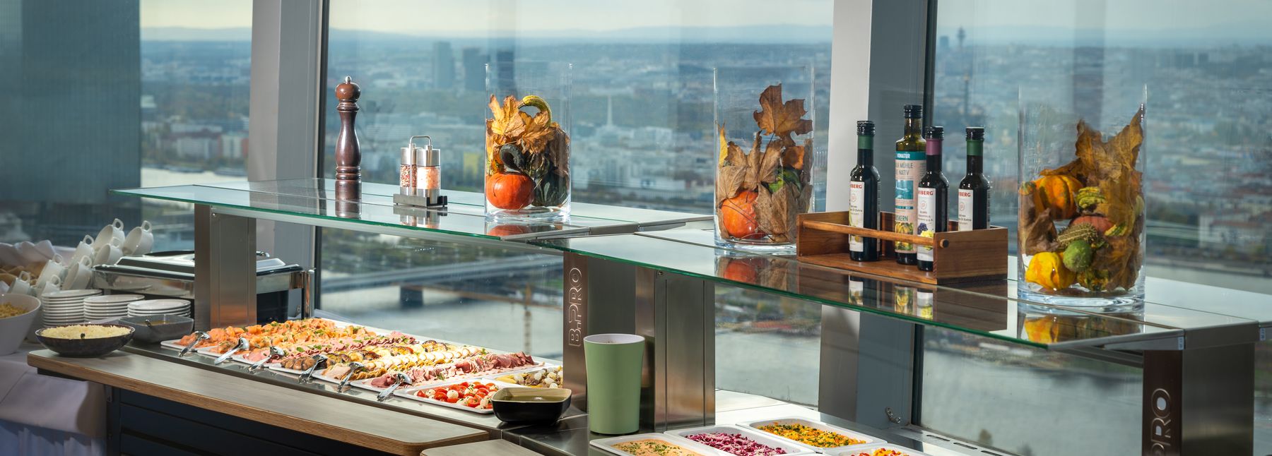 Brunchbuffet im Turmcafé des Donauturms in Wien mit einer Auswahl an Speisen auf einer Glasvitrine und Blick durch große Fenster über die Stadt.