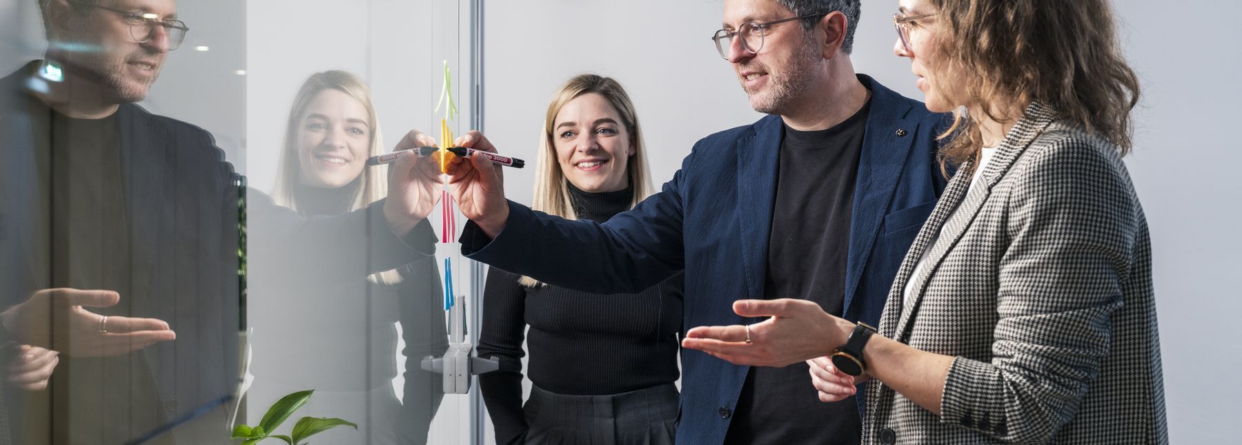 Drei Personen in einer Arbeitssituation. Zwei Frauen und ein Mann, der ein Post-it an eine Glaswand schreibt.