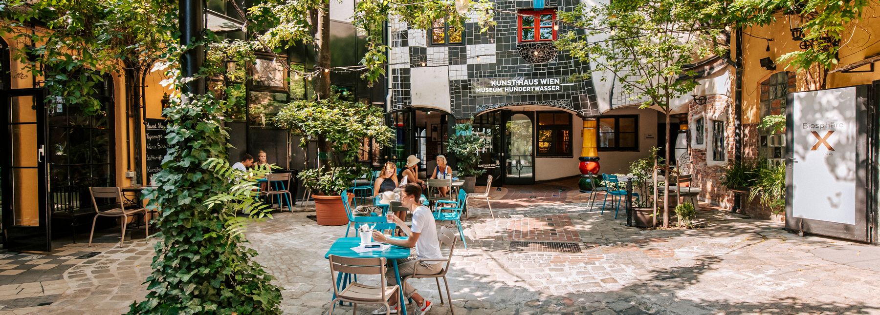 Blick in den begrünten Innenhof des KunstHausWien mit Kopfsteinpflaster, bunten Café-Tischen und Sitzgelegenheiten; zwischen Bäumen sitzen Gäste im Schatten, im Hintergrund die charakteristische Fassade des Museums mit farbigen Fenstern.