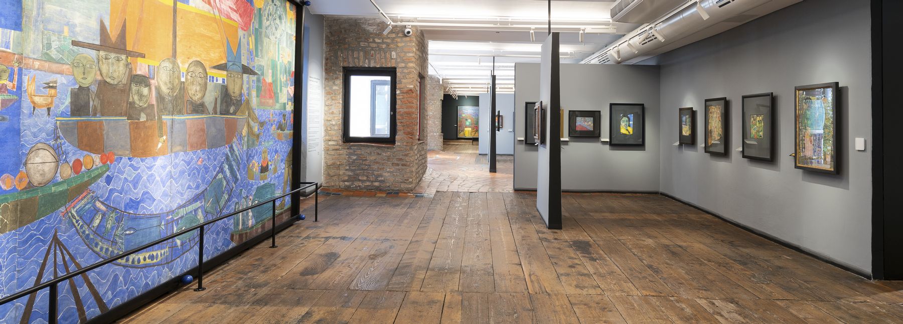 Ausstellungsraum im Museum Hundertwasser im KunstHausWien mit breitem Holzboden und moderner Beleuchtung. Links ein großformatiges, farbenreiches Wandbild, rechts mehrere gerahmte Kunstwerke an grauen Wänden sowie gemauerte Raumelemente.