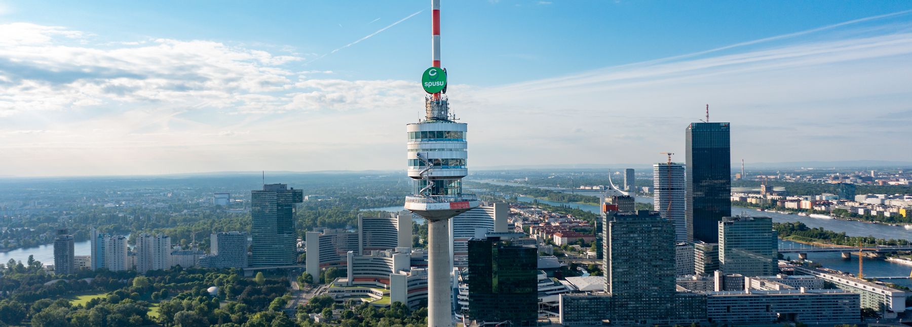 Blick aus der Luft auf den Donauturm in Wien mit rot-weißer Antennenspitze, umgeben von grünen Parkanlagen, moderner Hochhausarchitektur und der Donau unter blauem Himmel.
