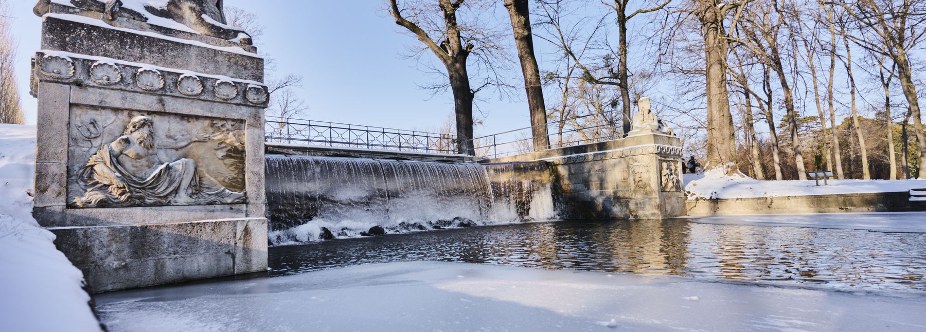 Die steinerne Kaskadenbrücke mit Statuen im Schlosspark Laxenburg, umgeben von einer winterlichen Landschaft mit teilweise gefrorenem Wasser und Schnee.