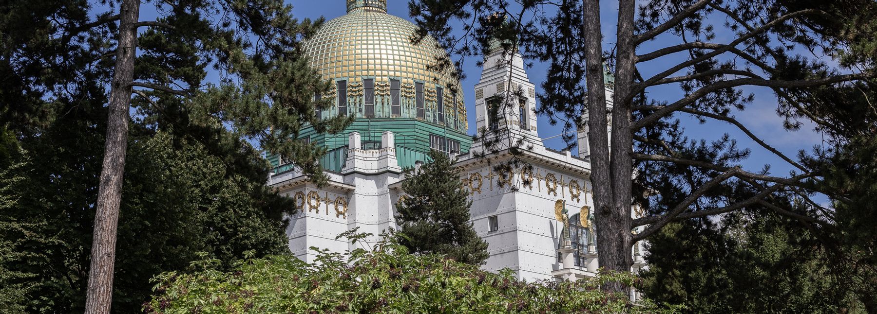 Otto-Wagner-Kirche am Steinhof, weiß mit oxidierten Kupferkuppeln und goldenen Details; im Vordergrund Bäume und Sträucher.