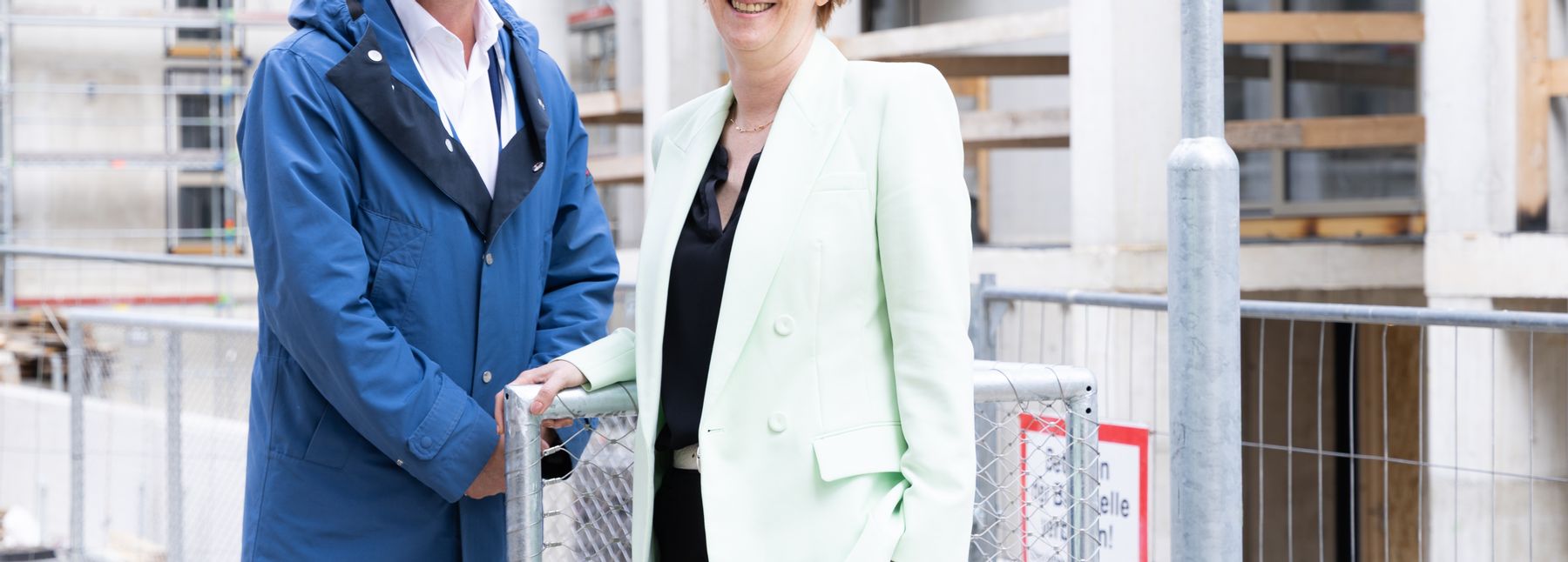 Ein Mann und eine Frau, die vor einem Betonbau stehen, lächeln in die Kamera