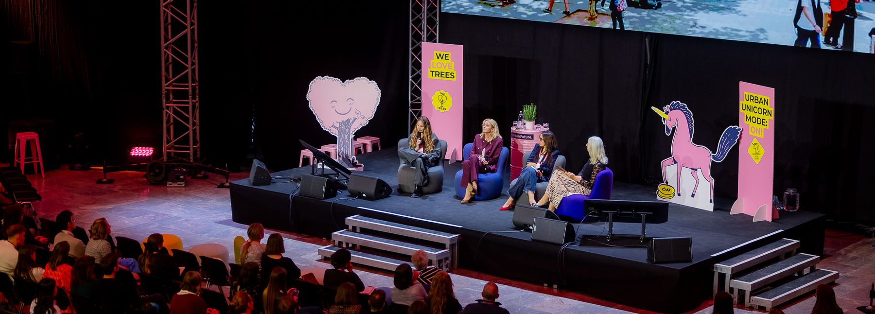 Ein Veranstaltungssaal mit Publikum und großer Leinwand, davor eine Gruppe von Menschen auf einer Bühne