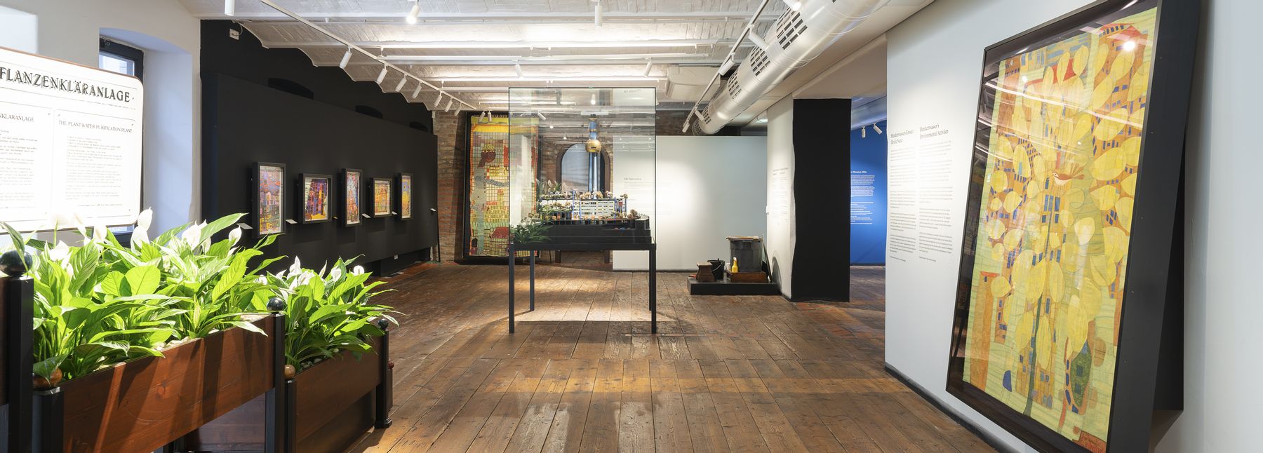 Heller Ausstellungsraum im Museum Hundertwasser im KunstHausWien mit Holzboden, weißer Gewölbedecke und Spotbeleuchtung; an den Wänden hängen farbenreiche Kunstwerke, in der Mitte stehen Vitrinen und Pflanzenelemente.