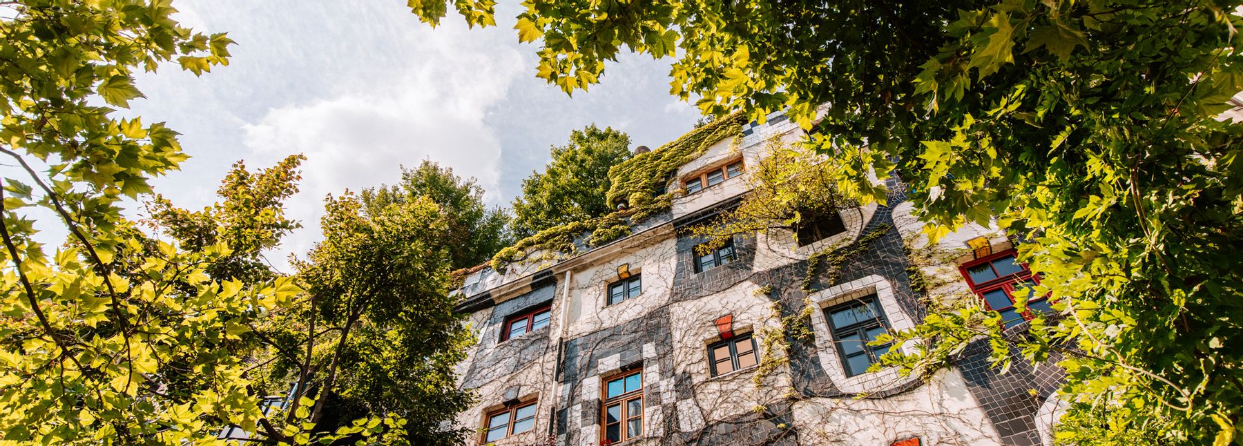 Froschperspektive auf die begrünte Fassade des KunstHausWien mit organisch geformter Architektur, bunten Fensterrahmen und bepflanztem Dach, eingerahmt von dichtem grünem Laub vor hellem Himmel.