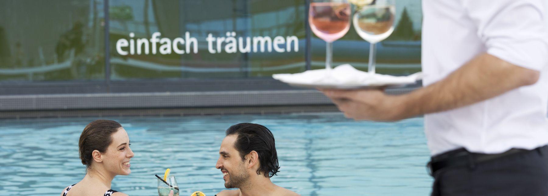 Ein Mann und eine Frau stehen im Außenbecken der Therme Wien und trinken Cocktails; am Rand des Beckens ist ein Kellner zu sehen, der zwei Cocktails auf einem Tablett trägt.