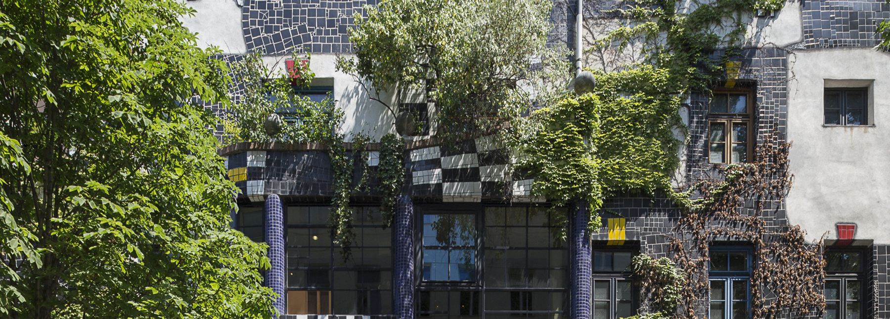 Frontalansicht der Fassade des KunstHausWien mit wellenförmiger Architektur, dunklen Keramikflächen, hellen Putzbereichen und farbigen Fensterrahmen; Pflanzen wachsen an der Fassade und auf dem Dach, im Vordergrund ein gepflasterter Platz mit einem Baum.