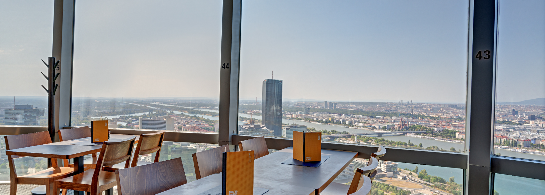 Innenansicht des Turmcafés im Donauturm in Wien mit Holztischen und großen Panoramafenstern mit Blick über die Wiener Skyline, die Donau und umliegende Grünflächen.