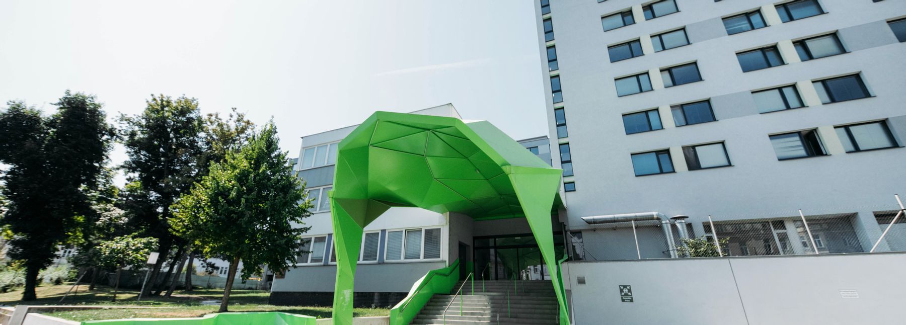 Eingangsbereich des Base 19-Gebäudes mit einer grünen Kunstinstallation vor dem kastenförmigen Bau.