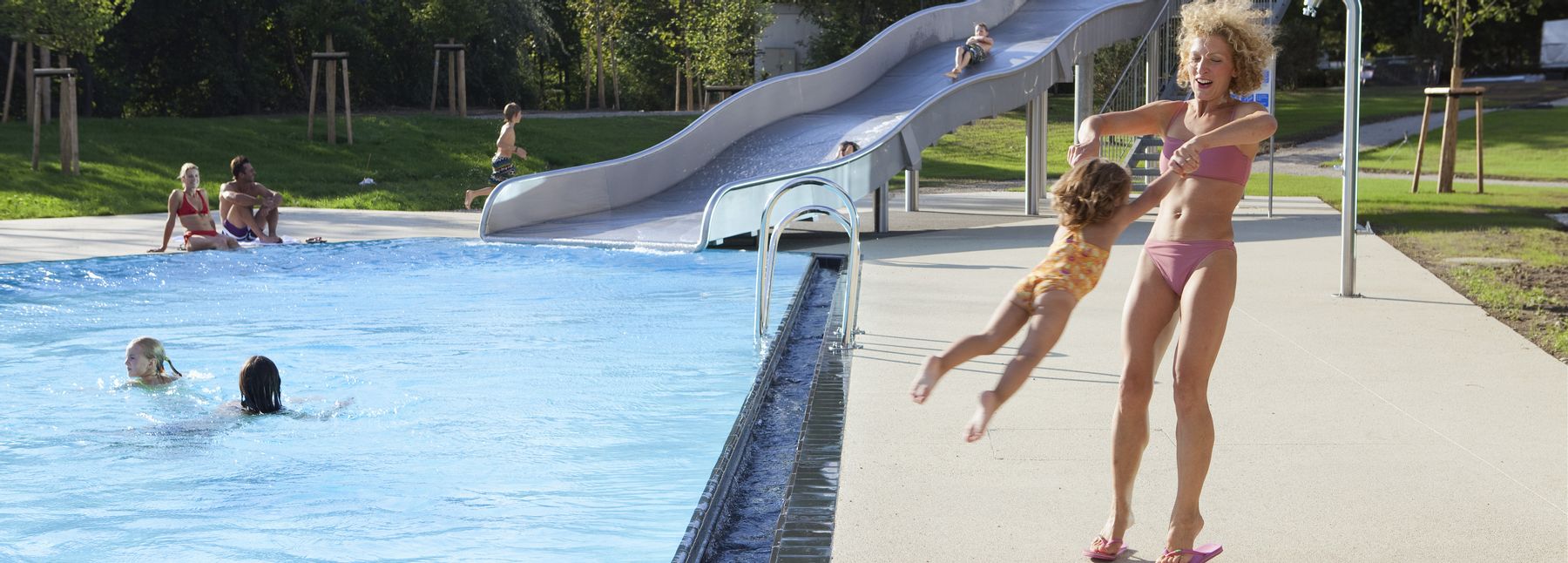 Außenbecken der Therme Wien mit einer Metallrutsche, auf der Kinder rutschen; im Vordergrund drehen sich eine Frau und ein Kind gemeinsam, während sie sich an den Händen halten.
