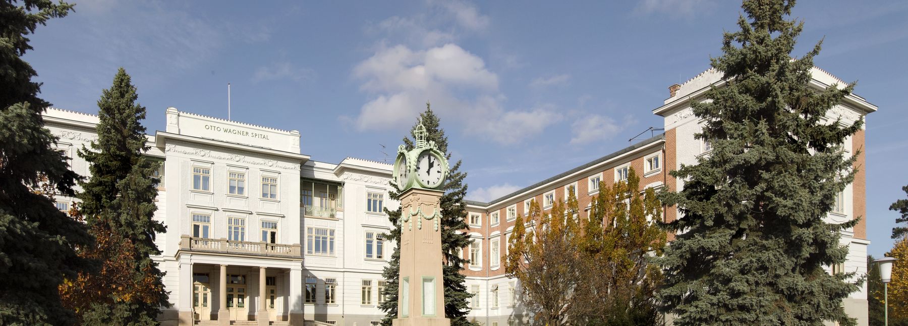 Direktionsgebäude am Otto-Wagner-Areal, weiß und teilweise aus Backstein; im Vordergrund eine Standuhr und Bäume.