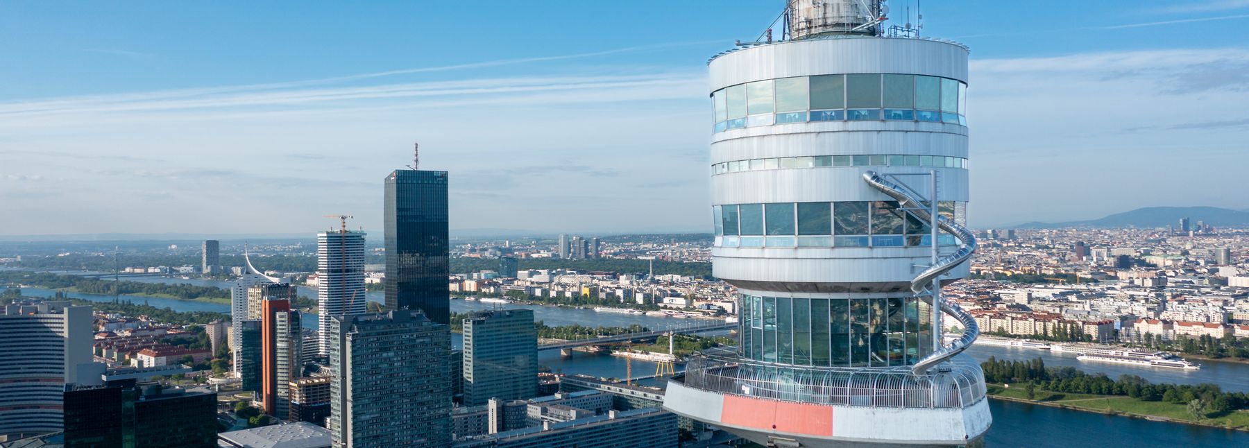 Oberer Bereich des Donauturms in Wien mit spiralförmiger Rutsche, im Hintergrund die Wiener Skyline, die Donau und grüne Parkanlagen unter klarem Himmel.