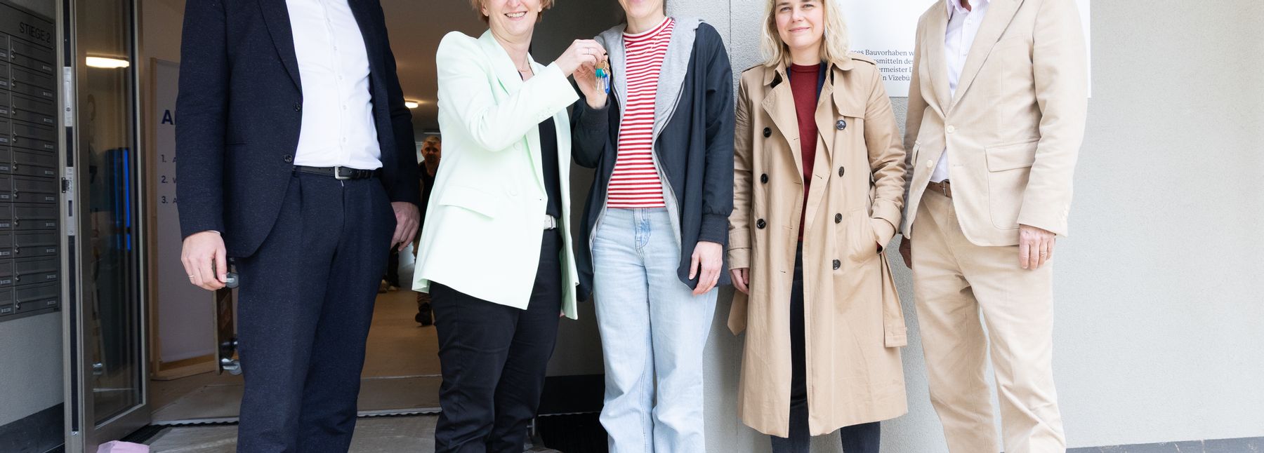 Eine Gruppe an Personen lächelt in die Kamera, eine Frau überreicht einer anderen Frau einen Schlüssel