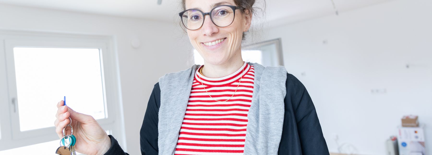 Eine Frau steht in einer leeren Wohnung, hält einen Schlüsselbund in der Hand und lächelt in die Kamera