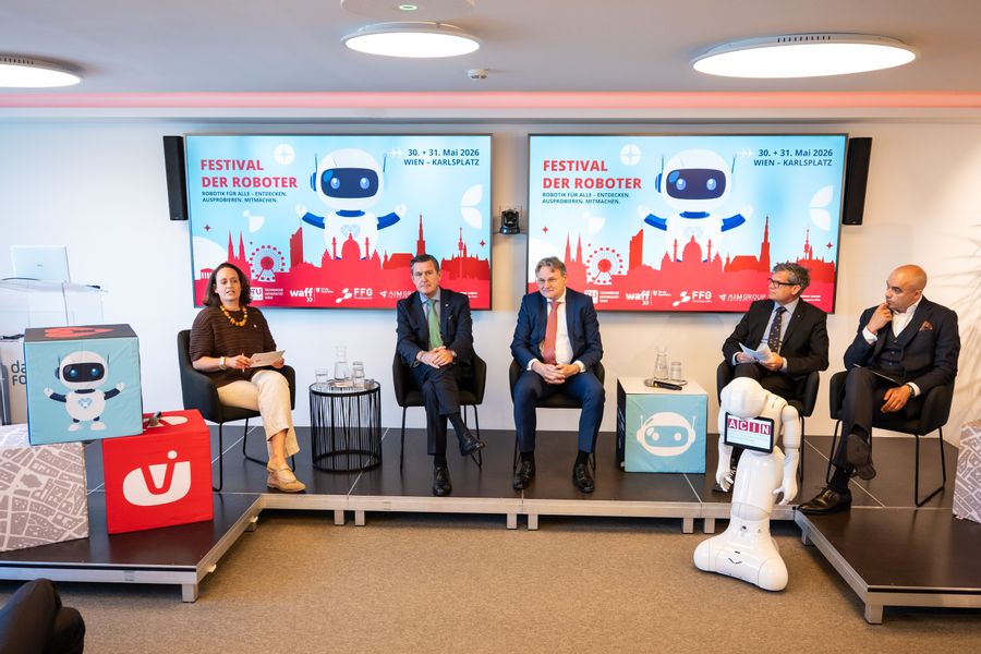 Gesprächsrunde aus vier Herren und einer Dame sitzend, im Hintergrund Bildschirme, im Vordergrund ein weißer kleiner Roboter