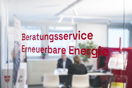 Durch eine Glasscheibe mit der roten Aufschrift „Beratungsservice Erneuerbare Energie“ sind zwei Personen unscharf bei einem Gespräch an einem Schreibtisch zu sehen.