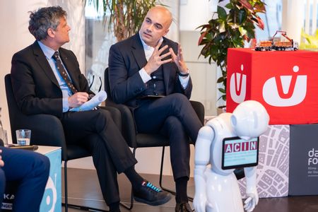 Zwei Herren bei einem Hintergrundgespräch, einer spricht, der andere hört zu, im Vordergrund steht ein weißer kleiner Roboter