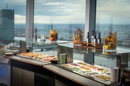 Brunchbuffet im Turmcafé des Donauturms in Wien mit einer Auswahl an Speisen auf einer Glasvitrine und Blick durch große Fenster über die Stadt.