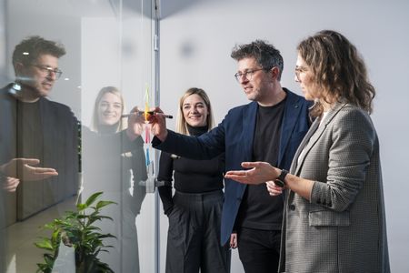 Drei Personen in einer Arbeitssituation. Zwei Frauen und ein Mann, der ein Post-it an eine Glaswand schreibt.