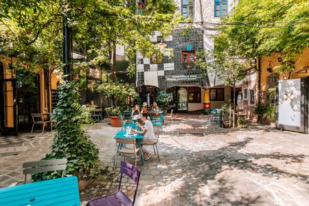 Blick in den begrünten Innenhof des KunstHausWien mit Kopfsteinpflaster, bunten Café-Tischen und Sitzgelegenheiten; zwischen Bäumen sitzen Gäste im Schatten, im Hintergrund die charakteristische Fassade des Museums mit farbigen Fenstern.