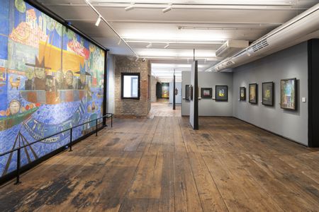 Ausstellungsraum im Museum Hundertwasser im KunstHausWien mit breitem Holzboden und moderner Beleuchtung. Links ein großformatiges, farbenreiches Wandbild, rechts mehrere gerahmte Kunstwerke an grauen Wänden sowie gemauerte Raumelemente.