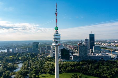 Blick aus der Luft auf den Donauturm in Wien mit rot-weißer Antennenspitze, umgeben von grünen Parkanlagen, moderner Hochhausarchitektur und der Donau unter blauem Himmel.