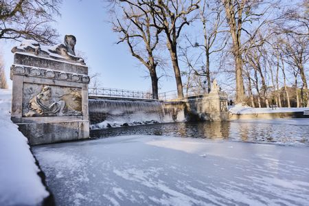 Die steinerne Kaskadenbrücke mit Statuen im Schlosspark Laxenburg, umgeben von einer winterlichen Landschaft mit teilweise gefrorenem Wasser und Schnee.