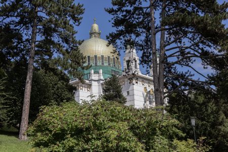 Otto-Wagner-Kirche am Steinhof, weiß mit oxidierten Kupferkuppeln und goldenen Details; im Vordergrund Bäume und Sträucher.