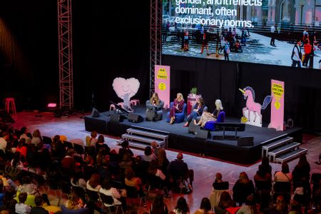 Ein Veranstaltungssaal mit Publikum und großer Leinwand, davor eine Gruppe von Menschen auf einer Bühne