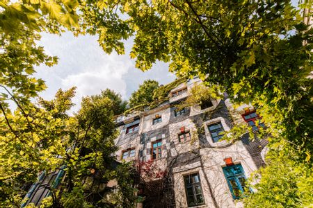 Froschperspektive auf die begrünte Fassade des KunstHausWien mit organisch geformter Architektur, bunten Fensterrahmen und bepflanztem Dach, eingerahmt von dichtem grünem Laub vor hellem Himmel.