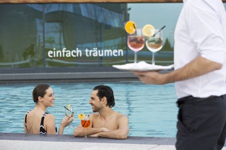 Ein Mann und eine Frau stehen im Außenbecken der Therme Wien und trinken Cocktails; am Rand des Beckens ist ein Kellner zu sehen, der zwei Cocktails auf einem Tablett trägt.