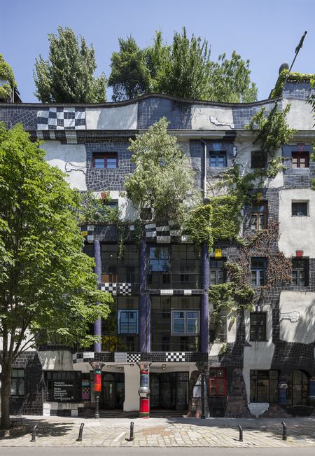 Frontalansicht der Fassade des KunstHausWien mit wellenförmiger Architektur, dunklen Keramikflächen, hellen Putzbereichen und farbigen Fensterrahmen; Pflanzen wachsen an der Fassade und auf dem Dach, im Vordergrund ein gepflasterter Platz mit einem Baum.