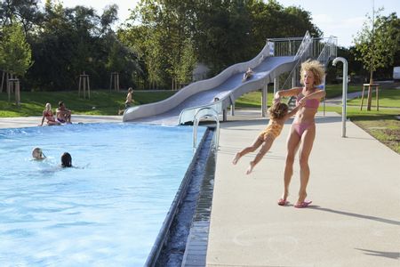 Außenbecken der Therme Wien mit einer Metallrutsche, auf der Kinder rutschen; im Vordergrund drehen sich eine Frau und ein Kind gemeinsam, während sie sich an den Händen halten.