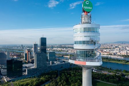 Oberer Bereich des Donauturms in Wien mit spiralförmiger Rutsche, im Hintergrund die Wiener Skyline, die Donau und grüne Parkanlagen unter klarem Himmel.