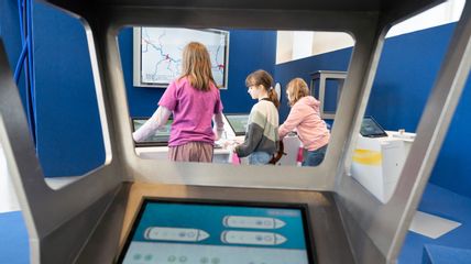 Im Zentrum des Bildes sind drei Mädchen im Volksschulalter zu sehen, die an interaktiven Bildschirmen der Ausstellung stehen.